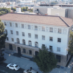 Aerial view of a four-story historic building with a red-tiled roof and white stucco exterior, located on a city block lined with trees and parked cars. The building features arched entrances, decorative details on the upper floors, and is surrounded by urban streets and nearby commercial structures in South Los Angeles.