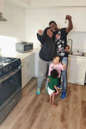 A smiling couple stands in their new kitchen holding up their house keys, with two young children at their feet. The family is standing on light wood flooring near a stove and sink, celebrating their move into a new home.
