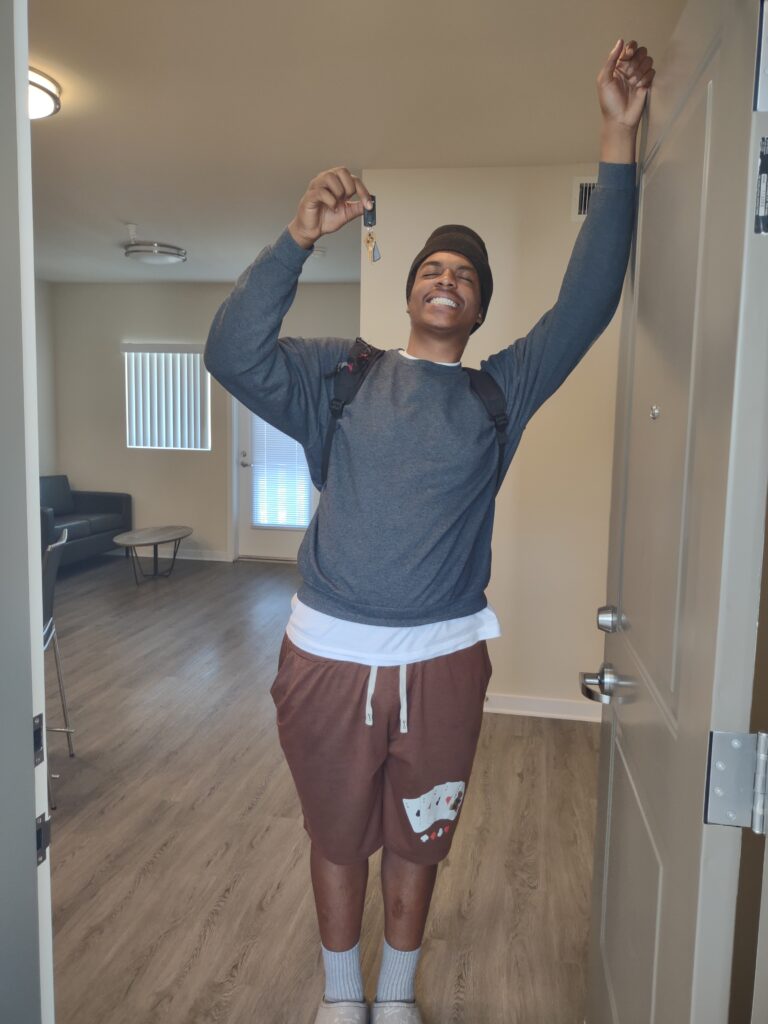 A young man stands smiling broadly in the doorway of his new apartment, holding up a set of keys in one hand and other raised, resting on the door. The apartment behind him has wood-style floors, modern furniture, and natural light coming through the windows.