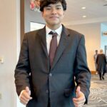 Smiling young man in a charcoal suit and burgundy tie gives two thumbs up in a bright lobby with patterned carpet. A success story of the the Workforce Development  for Youth and Job Pathways at CRCD