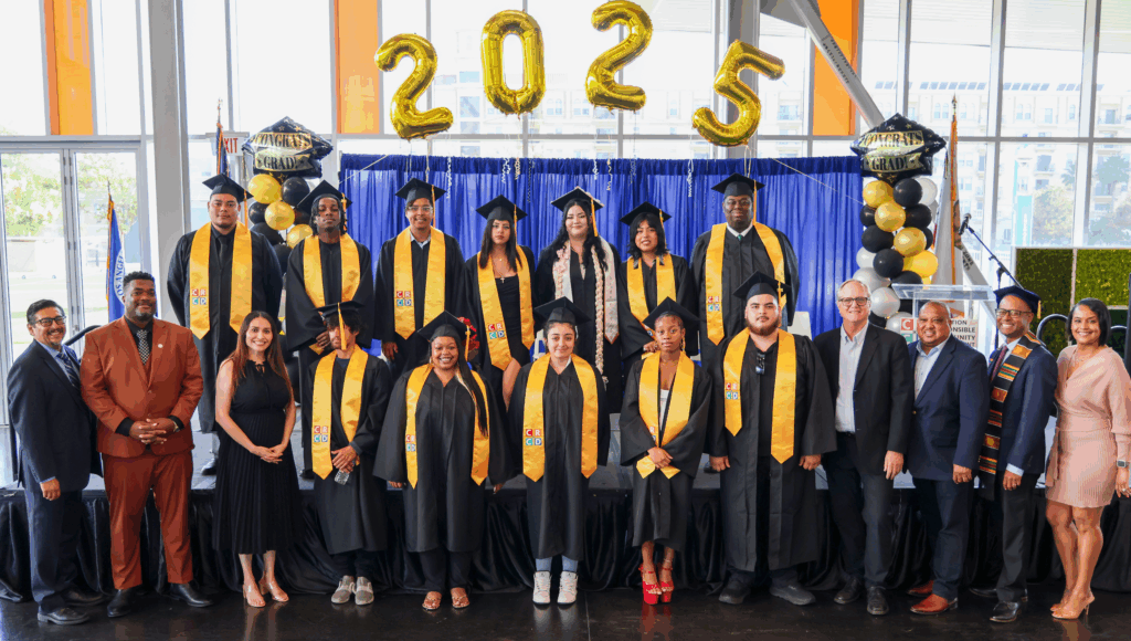 A group of CRCD Academy graduates from the Class of 2025 stand together wearing black caps and gowns with gold stoles featuring the CRCD logo. They are joined by CRCD staff and supporters in front of a blue curtain decorated with gold “2025” balloons and black, white, and gold balloons that say “Congrats Grad.” The setting is bright and celebratory, with large windows in the background letting in natural light. A success of the the Workforce Development  for Youth and Job Pathways at CRCD